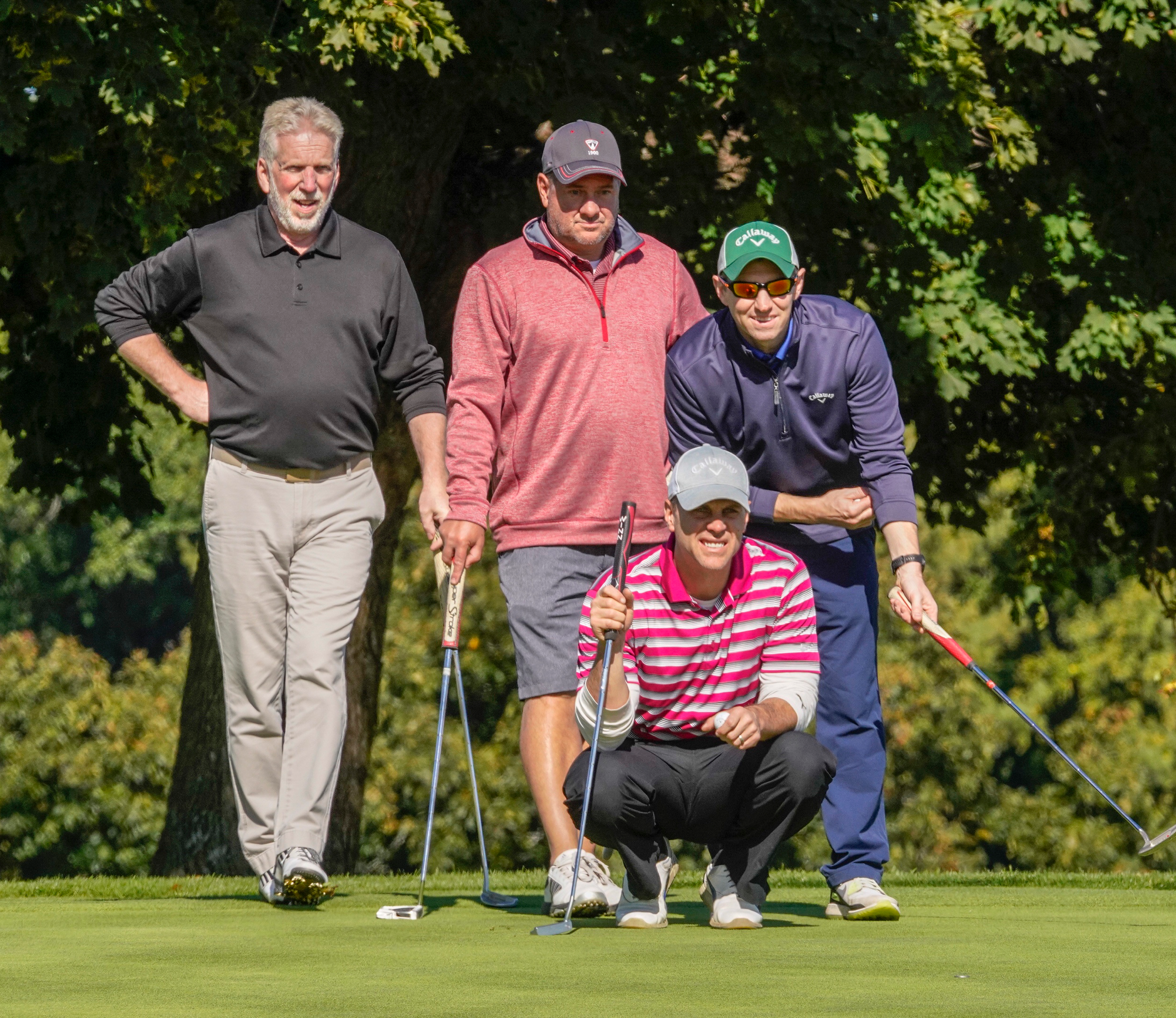 Four people stand on a golf green. One crouches, looking toward a cup.